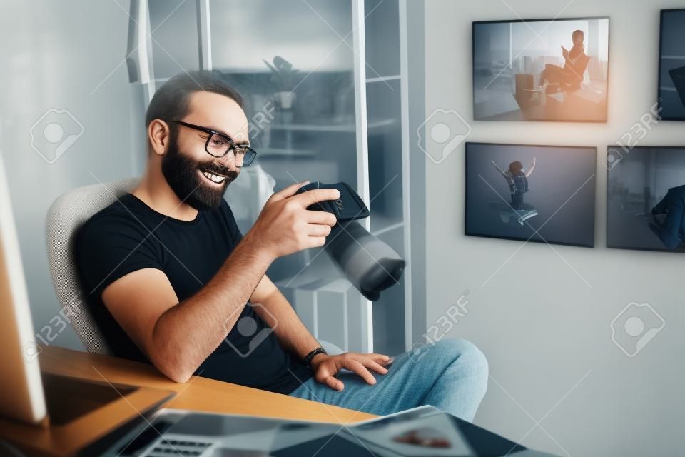 Retrato do homem barbudo sorridente que olha a tela do dispositivo digital enquanto senta-se na cadeira no apartamento.