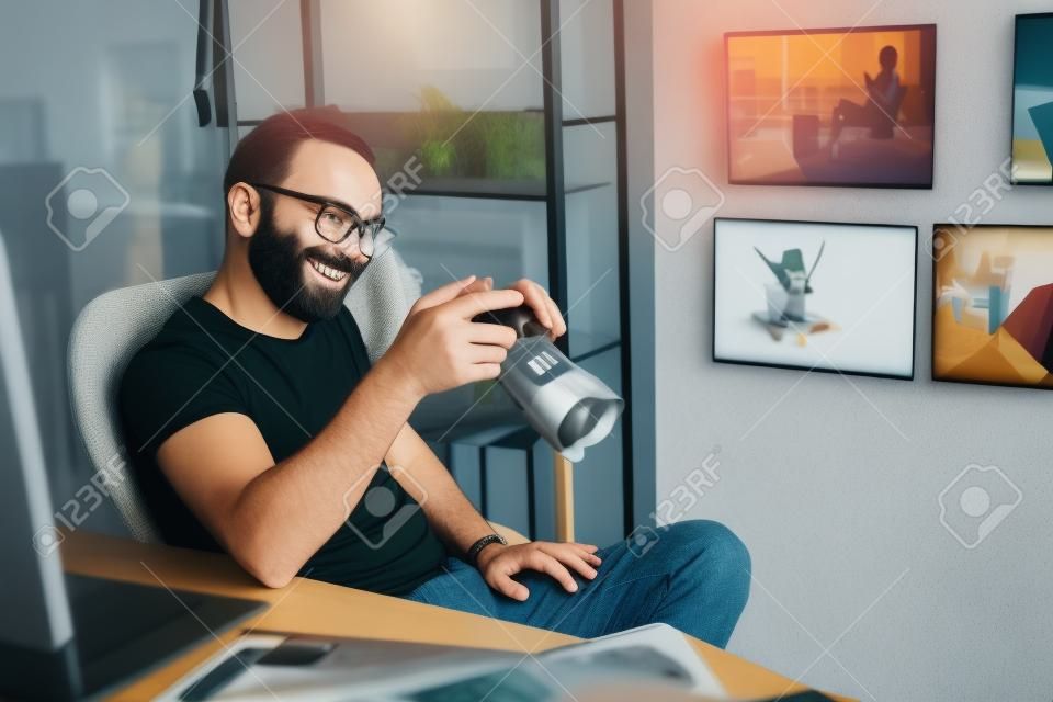 Retrato do homem barbudo sorridente que olha a tela do dispositivo digital enquanto senta-se na cadeira no apartamento.