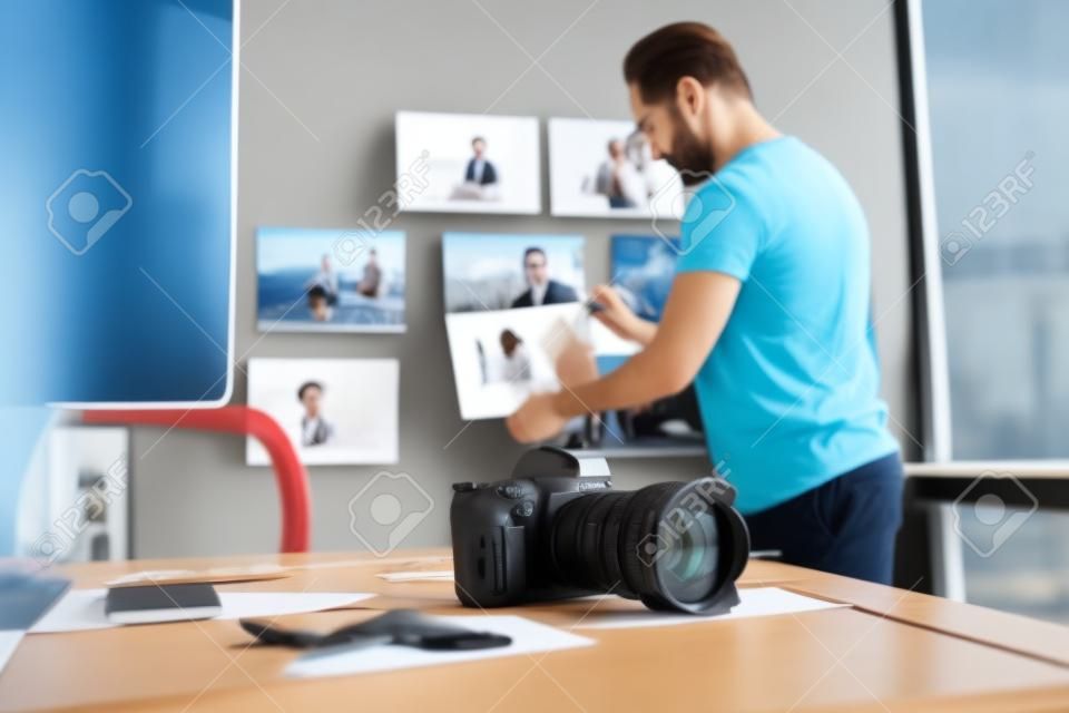 Focus on camera and photos locating on desk in office. Side view unshaven serene man putting pictures on background. Creativity and occupation concept