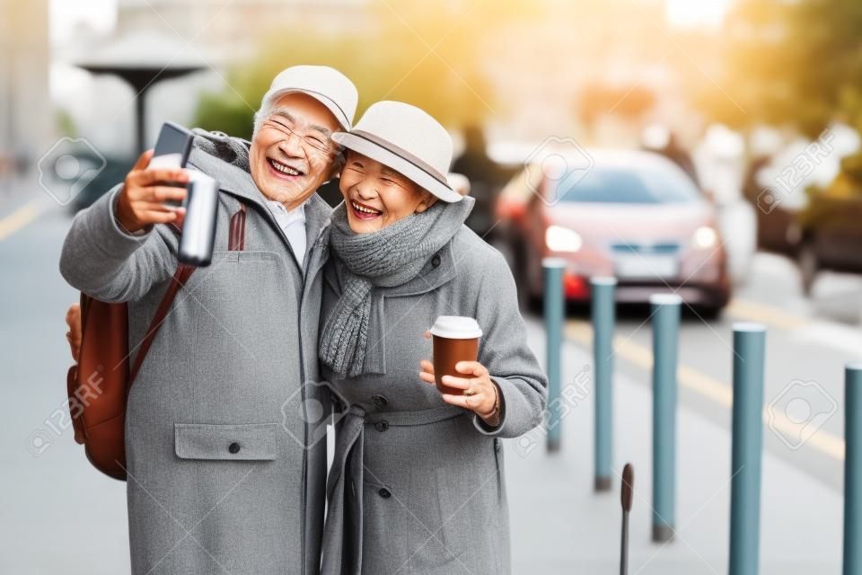 Süßer Moment. Zierliche ältere Frau mit Kaffee und ihrem grauhaarigen Ehemann nehmen selfie zusammen auf Straße. Sie lächeln und drücken Glück aus. Kopieren Sie Platz auf der rechten Seite