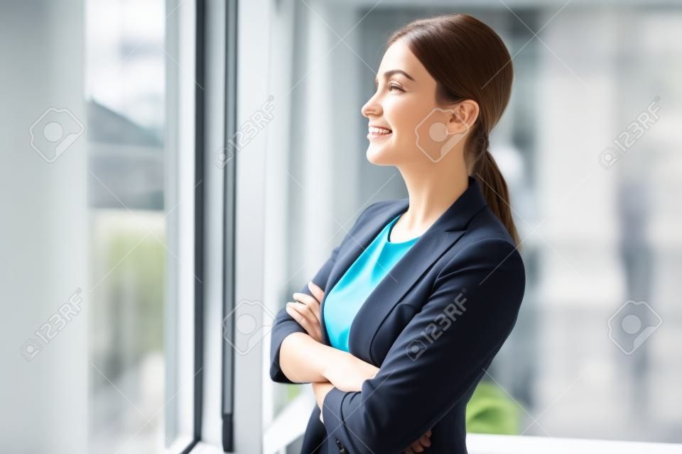 Side view smiling businesswoman watching at window while standing in room. Rest concept