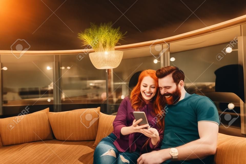 Excellent playlist. Smiling bearded man and lovely redhead lady sharing headphones and looking at mobile phone, They are sitting on the couch at outdoor cafe