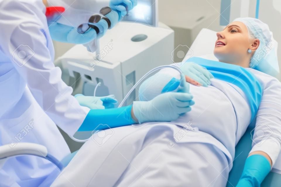 Esperando a gravidez. Médico em jaleco branco e luvas estéreis que examinam a mulher ruivo-haired com varredor do ultra-som
