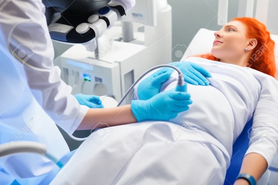 Spodziewając się ciąży. Lekarz w białym fartuchu laboratoryjnym i sterylnych rękawiczkach badający rudowłosą kobietę za pomocą ultrasonografu