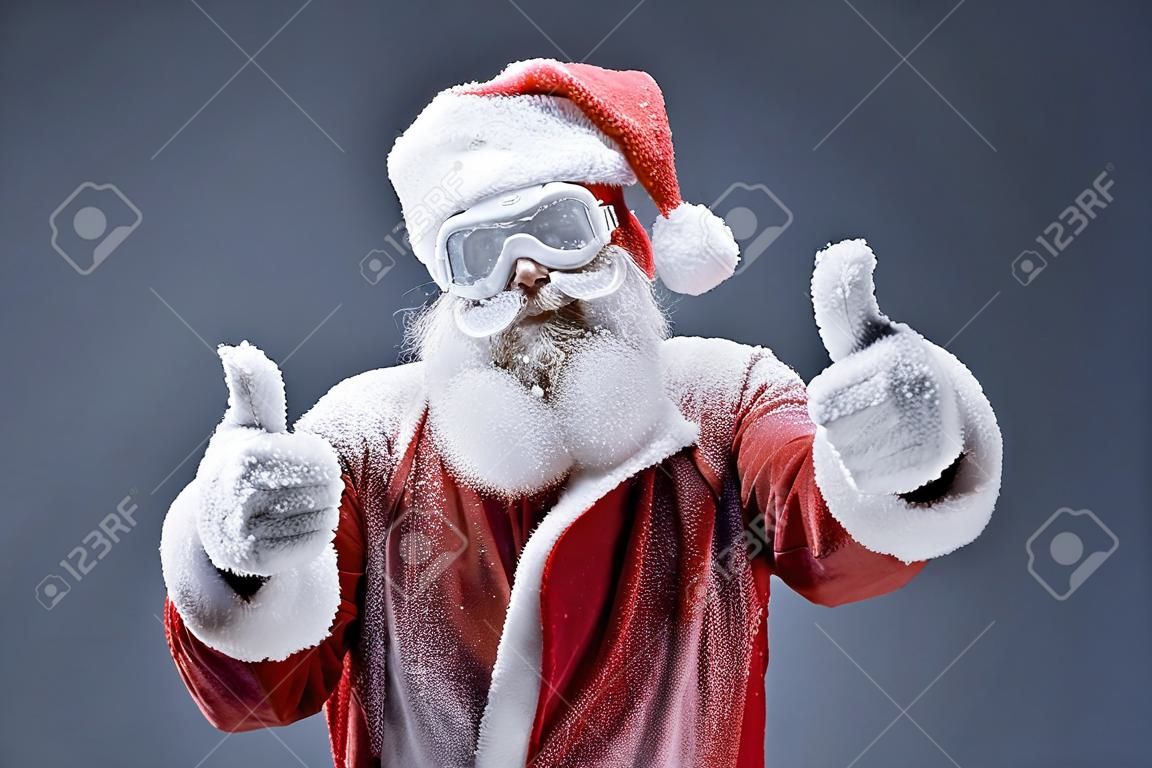 Portrait of bearded old man in Santa costume covered with snow showing thumbs up sign. Isolated on gray-blue background