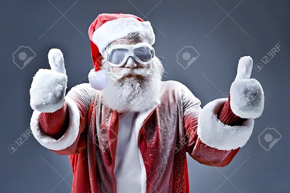 Retrato do homem velho barbudo no traje de Papai Noel coberto com neve que mostra os polegares acima do sinal. Isolado no fundo cinzento-azul