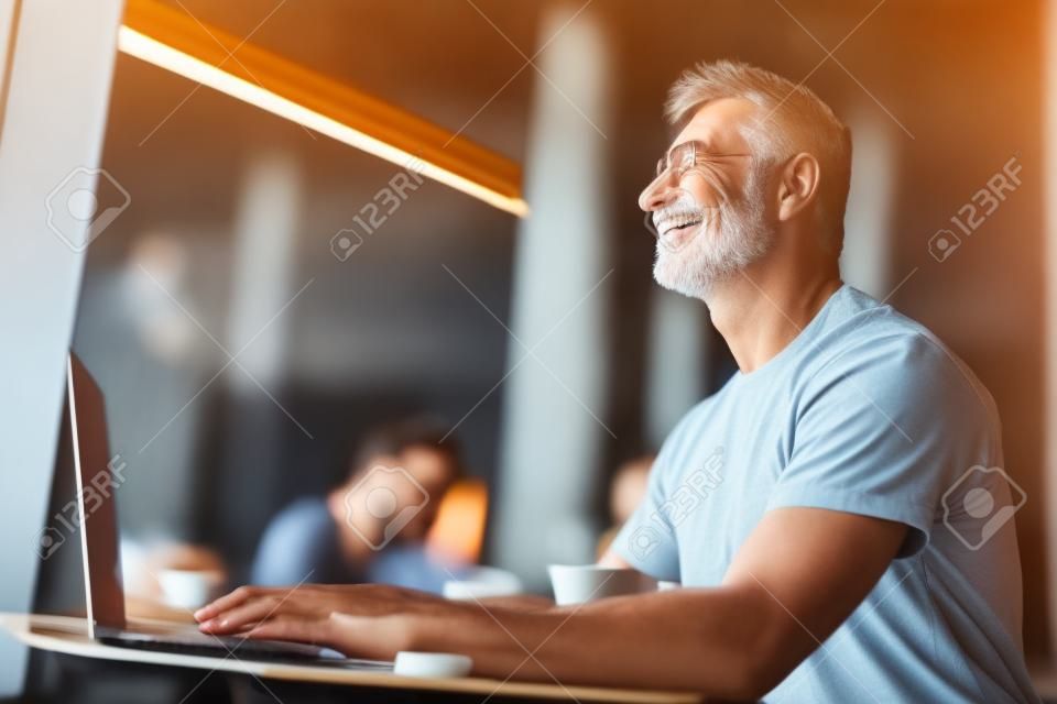 カフェに座っている魅力的な成熟した男。彼はラップトップのキーボードに横たわっている彼の手を見上げて微笑む