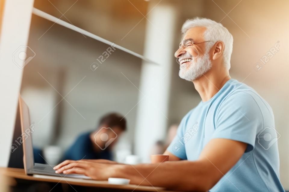 カフェに座っている魅力的な成熟した男。彼はラップトップのキーボードに横たわっている彼の手を見上げて微笑む