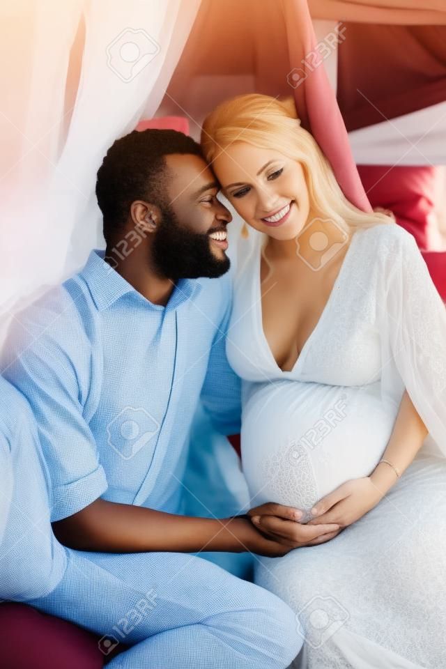 赤ちゃんを期待している異人種間のカップル。妊娠中の白人女性と彼女のアフリカの夫はお互いの腕の中で天蓋の下に座っています。
