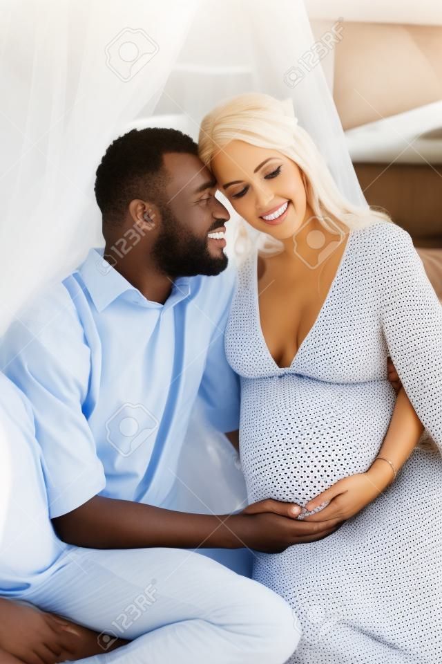 Coppia interrazziale in attesa del bambino. Donna caucasica incinta e suo marito africano seduti sotto il baldacchino l'uno nelle braccia dell'altro.
