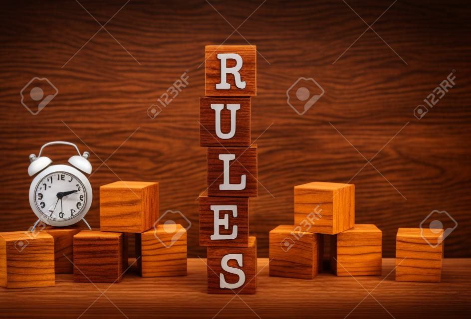 Five wooden cubes stacked vertically to form the word RULES on a brown background. Cubes are scattered nearby and there is a clock. Front View Concepts