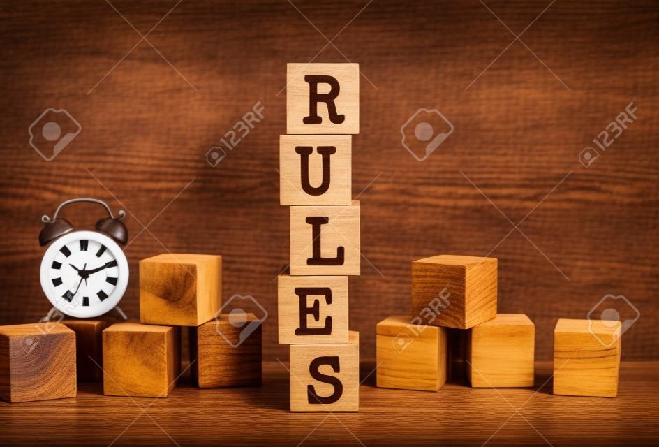 Five wooden cubes stacked vertically to form the word RULES on a brown background. Cubes are scattered nearby and there is a clock. Front View Concepts