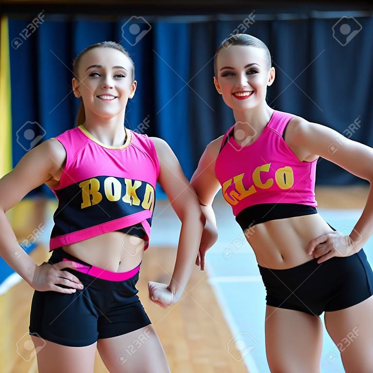 girls with fit body demonstrating muscles, smiling beautiful women in black and pink sportswear looking at camera, cheerleaders working out in sports club, cheerleaders smiling and posing at camera