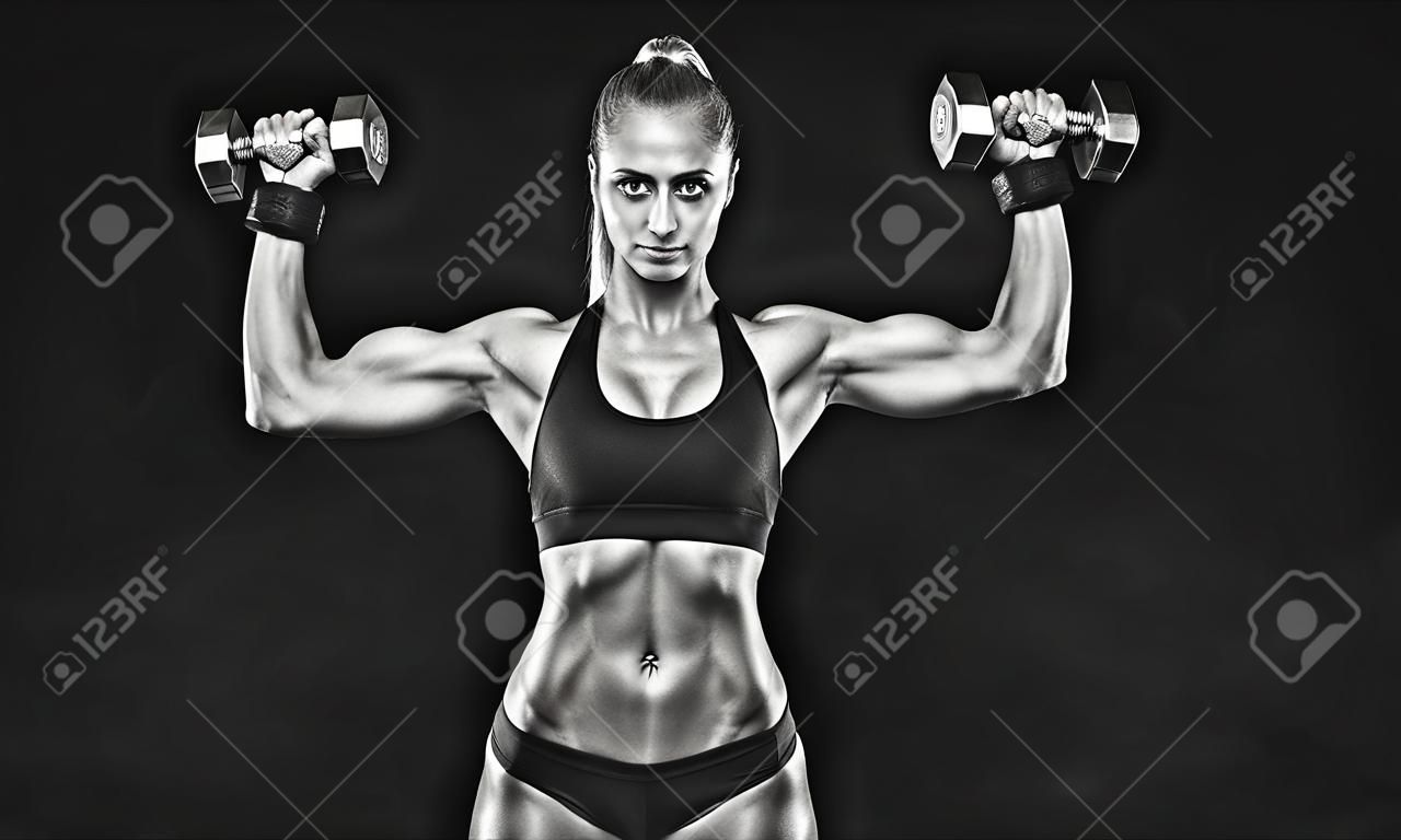 Black and white shot of young female bodybuilder working out with hand weights and shorts curling dumbbell over black background Caucasian woman with muscular body, abs Mockup.