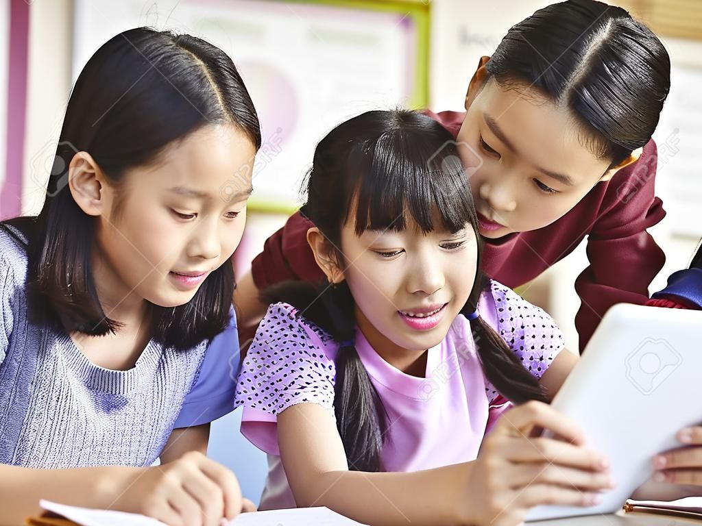 three asian elementary school girls friends looking at a tablet together during break in classroom.