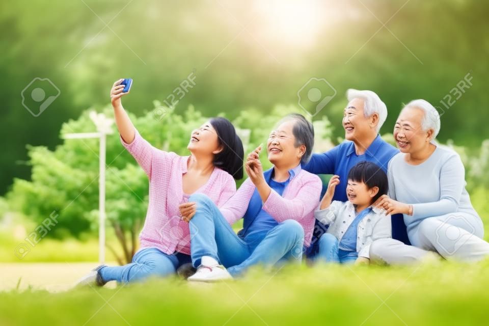 three generation happy asian family sitting on grass taking a selfie using mobile phone outdoors in park