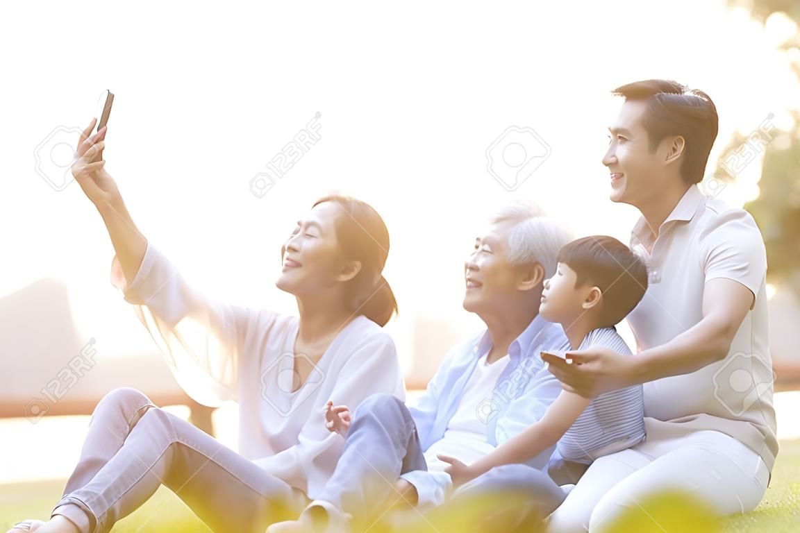 Drei Generationen glückliche asiatische Familie, die auf Gras sitzt und ein Selfie mit dem Handy im Freien im Park macht