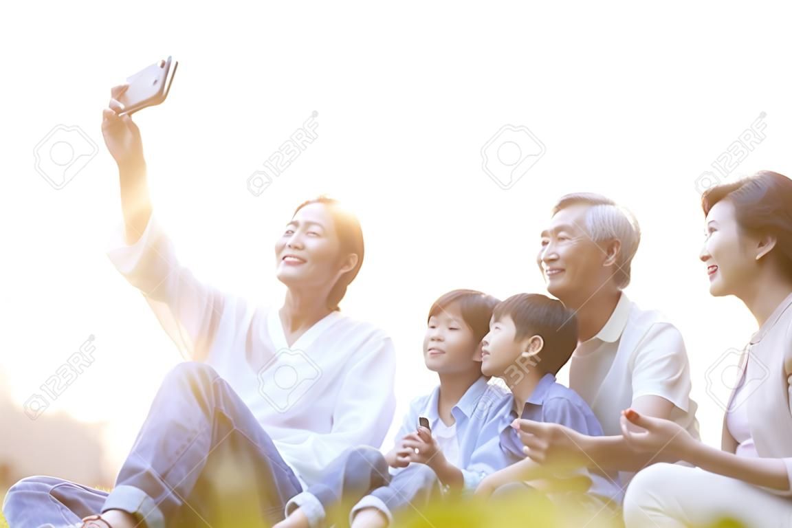 Drei Generationen glückliche asiatische Familie, die auf Gras sitzt und ein Selfie mit dem Handy im Freien im Park macht