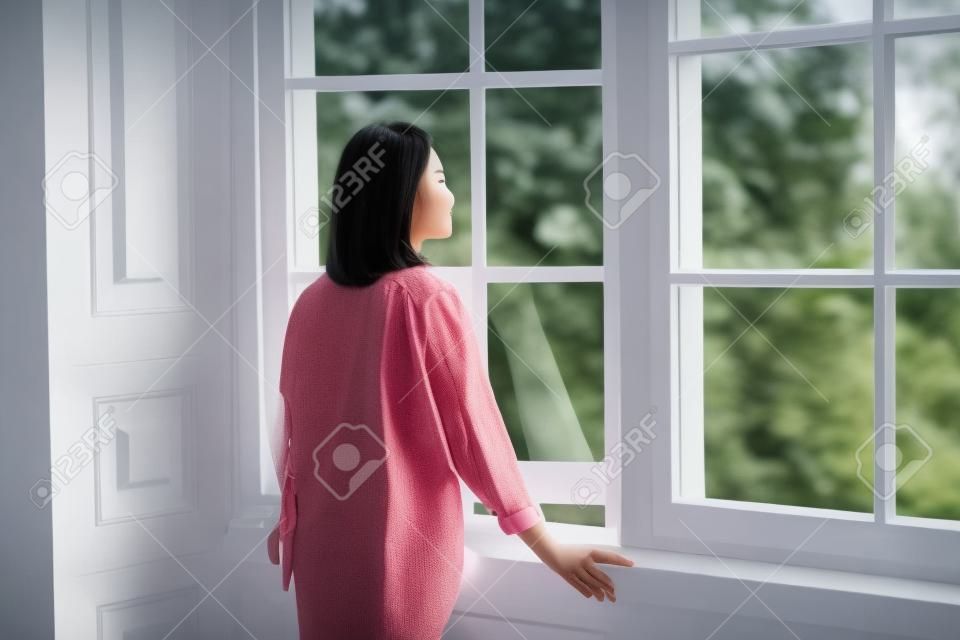 rear side view of a young asian woman looking out of window at home