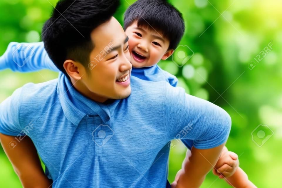 Młody azjatycki ojciec niosący syna na plecach, dobrze się bawiąc, ciesząc się przyrodą na świeżym powietrzu w parku