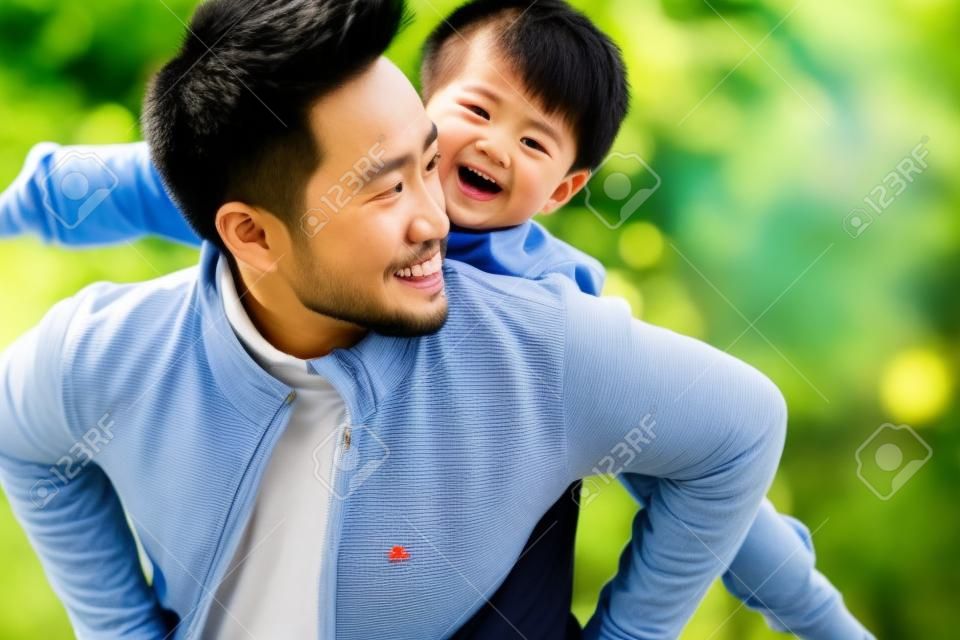 Młody azjatycki ojciec niosący syna na plecach, dobrze się bawiąc, ciesząc się przyrodą na świeżym powietrzu w parku
