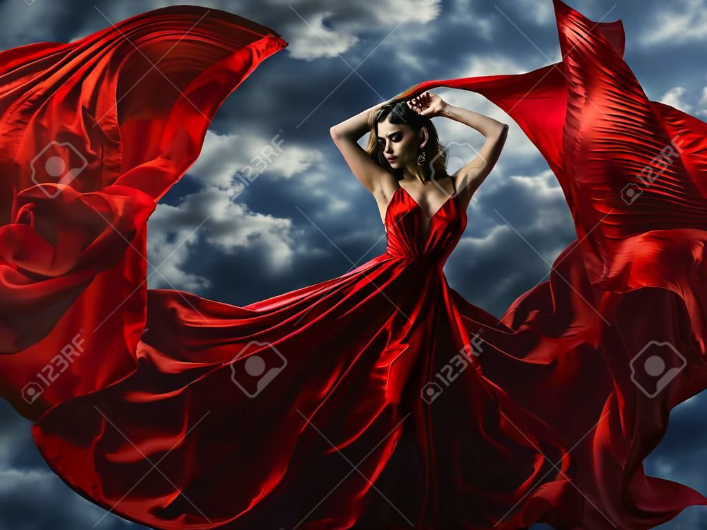 Woman in red evening dress, waving gown with flying long fabric over artistic sky background