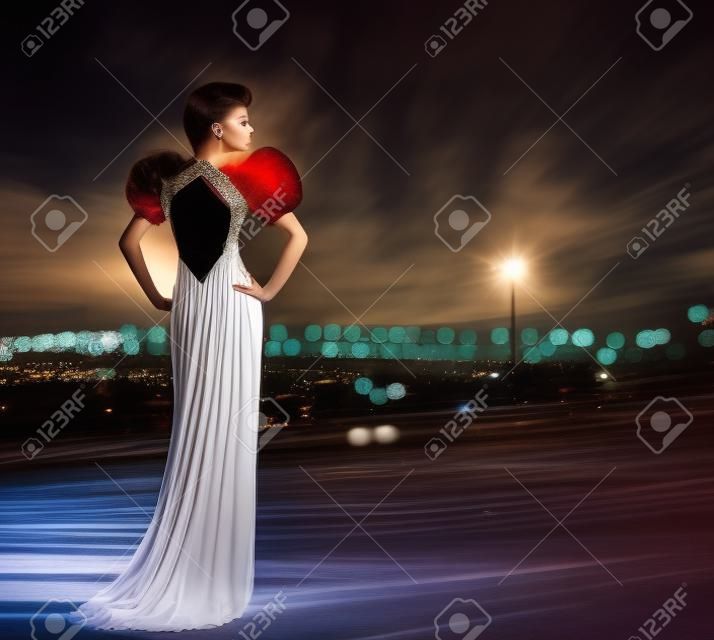 Vestido de noite de mulher Posing em luzes da noite da cidade, modelo de moda em vestido longo, vista traseira olhando para o lado, vida noturna urbana
