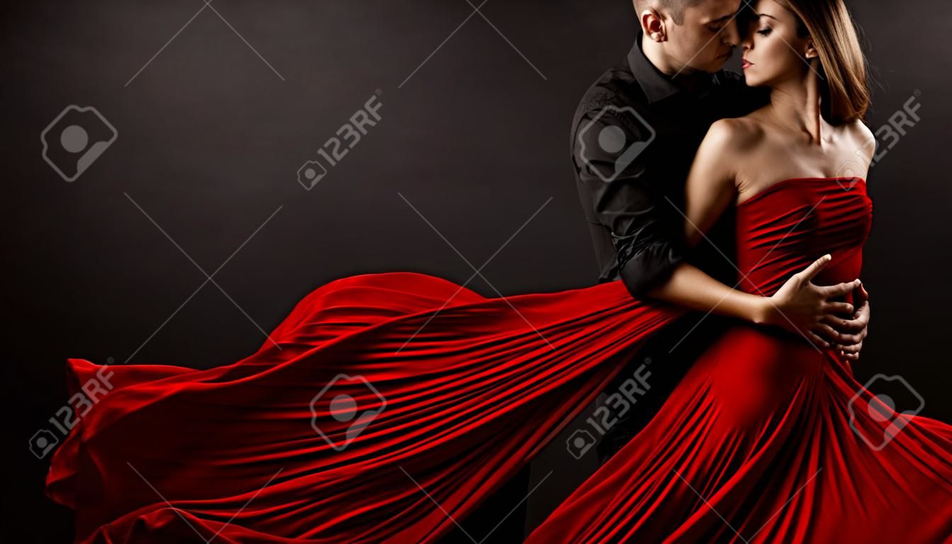 Coppia di amanti romantici che ballano. Uomo che abbraccia e bacia bella donna in abito volante rosso di seta. Ritratto di moda. Sfondo nero da studio