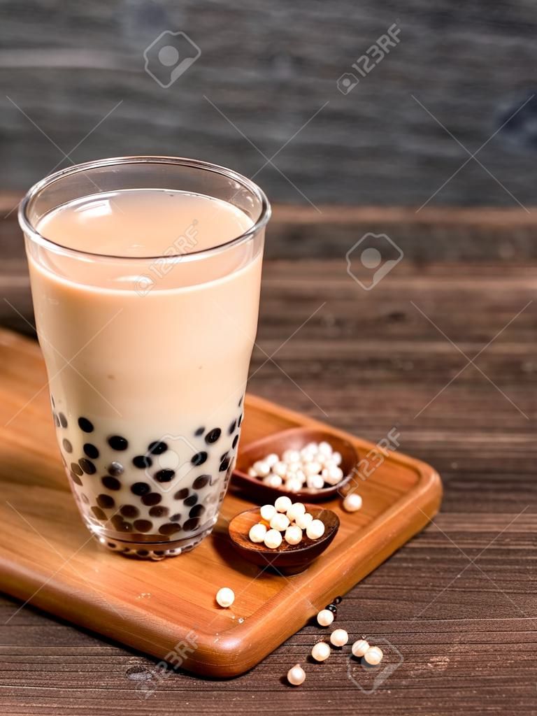 Köstlicher Schaummilchtee mit Tapiokaperlenkugel in Glas auf Holztisch und dunkelgrauem Ziegelsteinhintergrund, beliebtes Essen und Trinken in Taiwan, Nahaufnahme