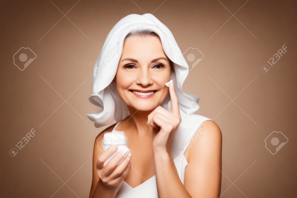 Smiling 50s middle aged woman putting facial cream on face looking at camera.