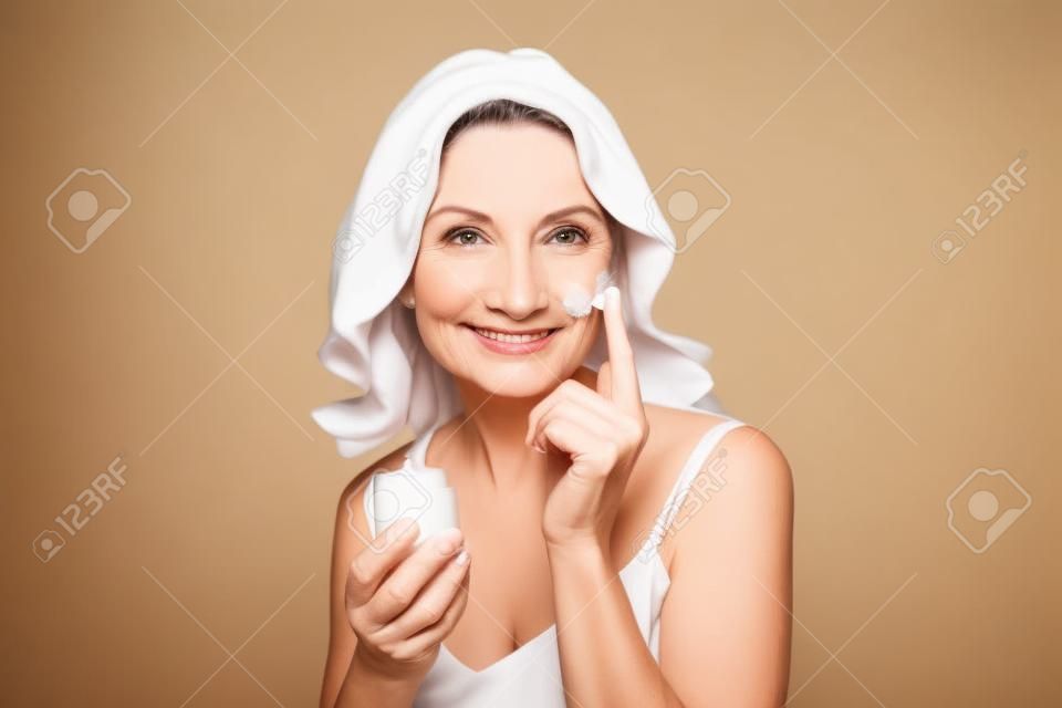Smiling 50s middle aged woman putting facial cream on face looking at camera.