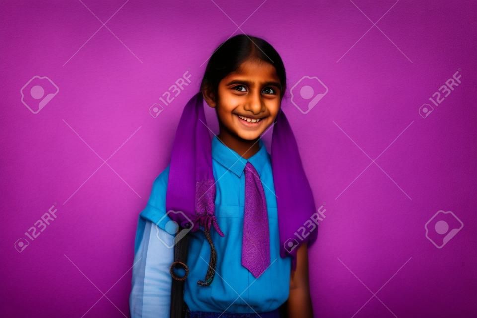 Portrait of indian schoolgirl with bag standing isolated on violet background.
