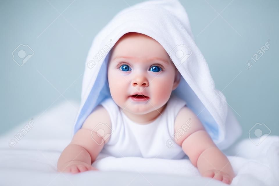 Cute baby girl or boy after shower with towel on head in white sunny bedroom. Child with big blue eyes relaxing in bed after bath