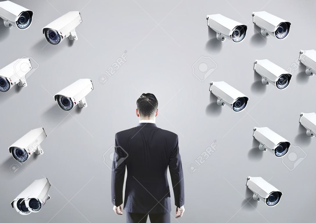 Catorze câmeras de CCTV penduradas em fileiras em uma parede cinza de frente para um homem de negócios que olha para a parede. Conceito de vigilância e monitoramento.