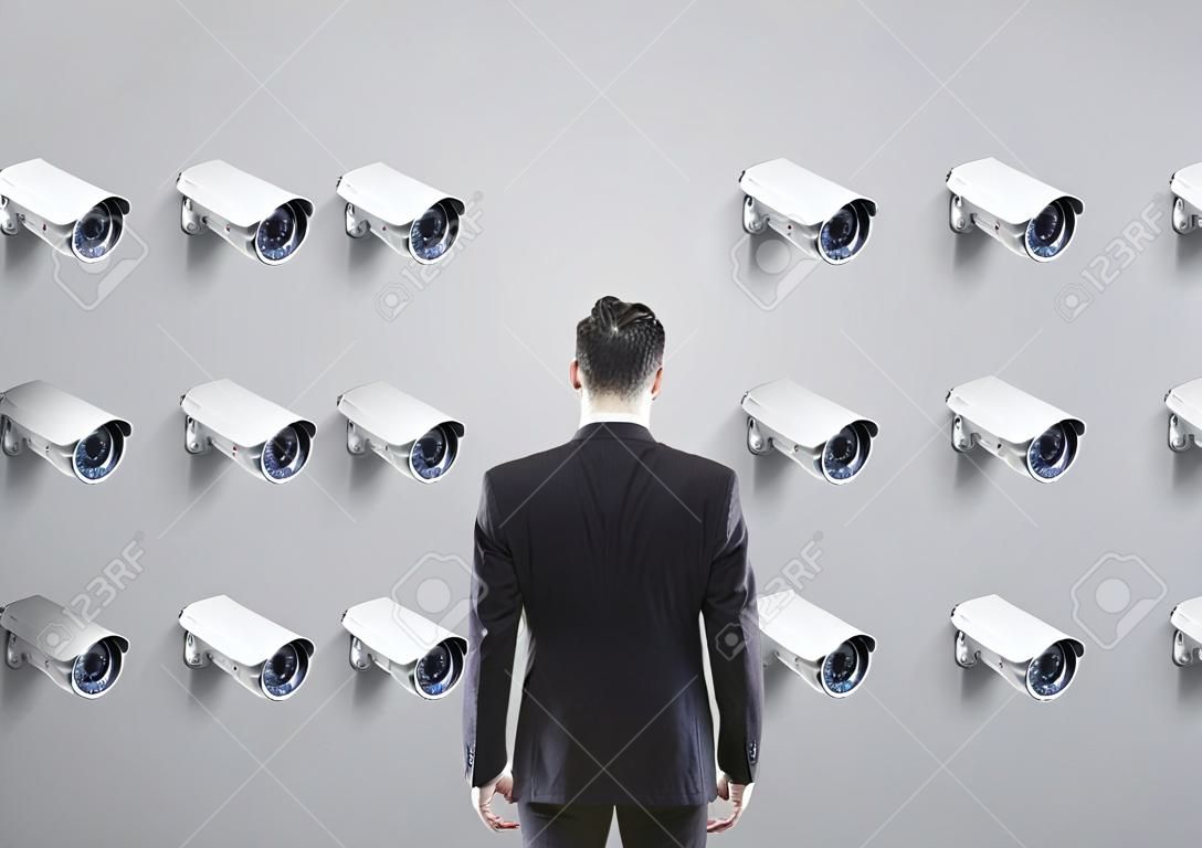 Fourteen CCTV cameras hanging in rows on one gray wall facing a businessman looking at the wall. Concept of surveillance and monitoring. Mock up