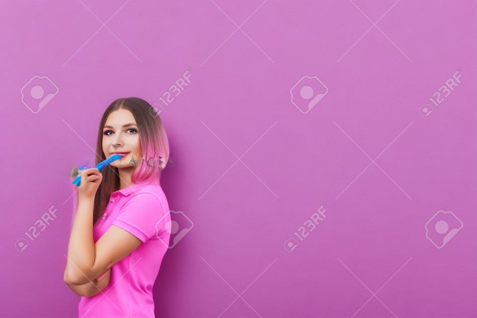 Portrait of young caucasian woman with long fair hair wearing pink polo shirt, holding marker and thinking standing near gray wall. Concept of planning. Mock up