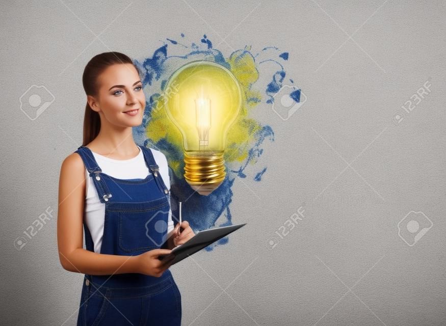 Atrakcyjna europejska młoda kobieta w drelichowym kombinezonie trzymająca planistę i zastanawiająca się przy betonowej ścianie z narysowanym na niej szkicem żarówki kolorowy pomysł na biznes. pojęcie kreatywności i burzy mózgów