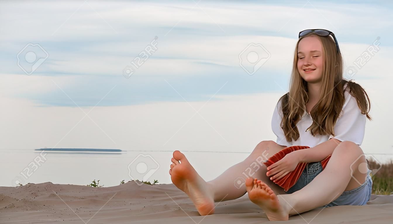 Одна красивая девочка-подросток с каштановыми волосами на улице в прекрасный летний день. Девушка делает себе массаж ступней. Крем для втирания от солнечных ожогов. На ступнях видны солнечные ожоги.