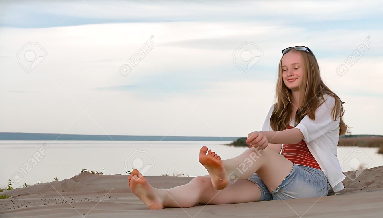Одна красивая девочка-подросток с каштановыми волосами на улице в прекрасный летний день. Девушка делает себе массаж ступней. Крем для втирания от солнечных ожогов. На ступнях видны солнечные ожоги.