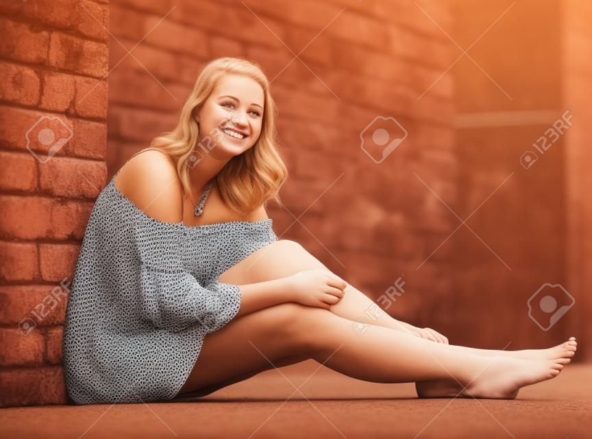 Joyeuse fille assise pieds nus près du mur de briques en robe à l'extérieur