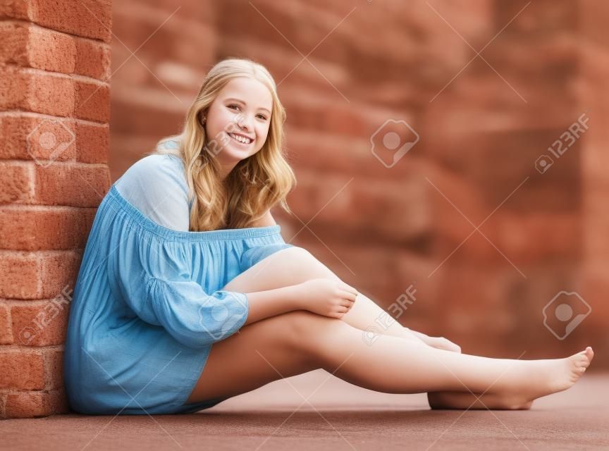 屋外のドレスでレンガの壁の近くに裸足で座っている陽気な女の子