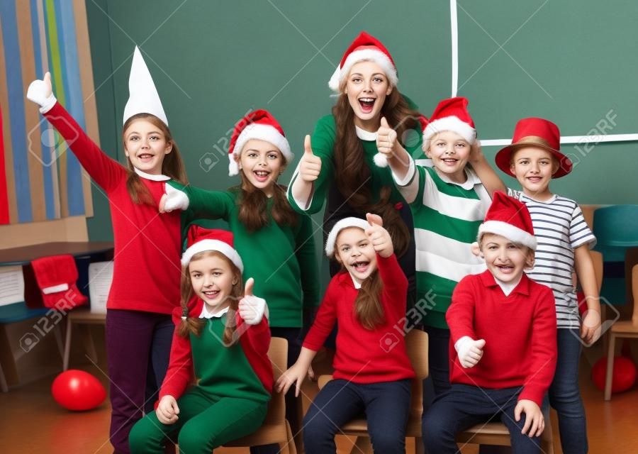 Grupo alegre de alunos com professora vestindo chapéus de Papai Noel posando juntos na sala de aula, dando polegares para cima