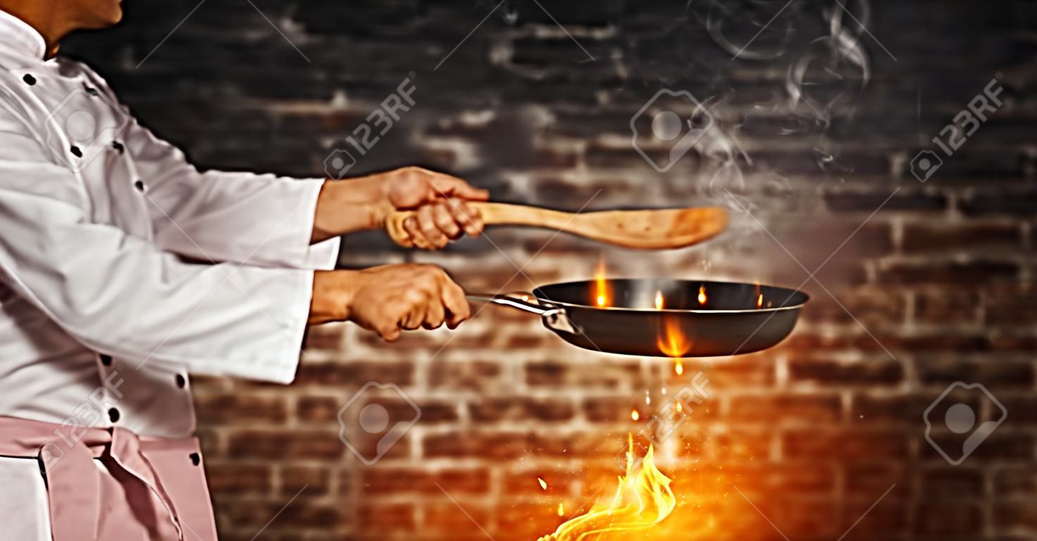 Close-up van chef-kok klaar om te koken, houden lege grill pan, vliegende beweging effect. Klaar voor productplaatsing. Oude baksteen muur op achtergrond