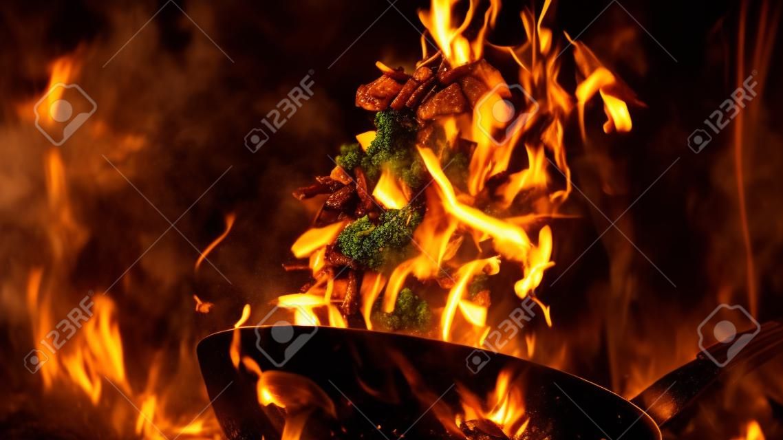 Closeup of chef throwing vegetable mix from wok pan in fire. Fresh asian food preparation on dark background.