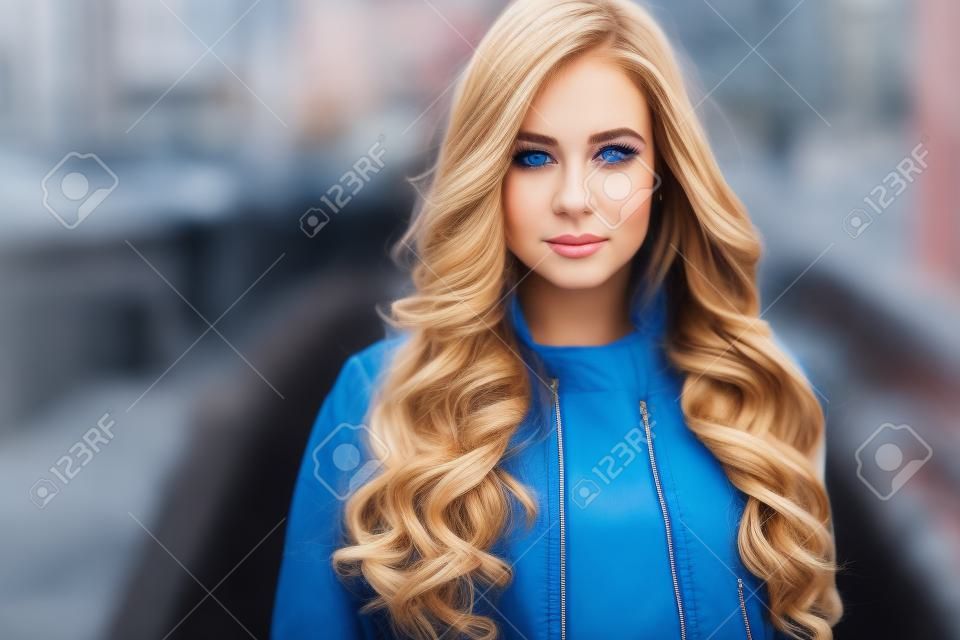 Retrato do close-up da jovem loira com lindos olhos azuis, vestindo jaqueta preta ao ar livre. Mulher bonita russa com penteado longo cabelo ondulado. Mulher em meio urbano.
