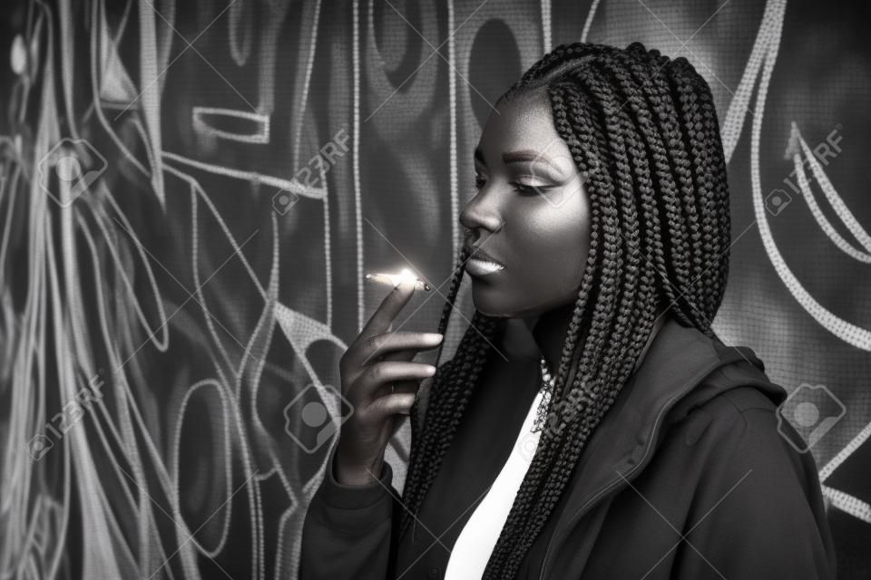 Geflochtene junge Frau raucht im Freien. Mit afrikanischen Zöpfen, durchbohrte Mode Frau. Umwandlung in Schwarz und Weiß, Getreide hinzugefügt.