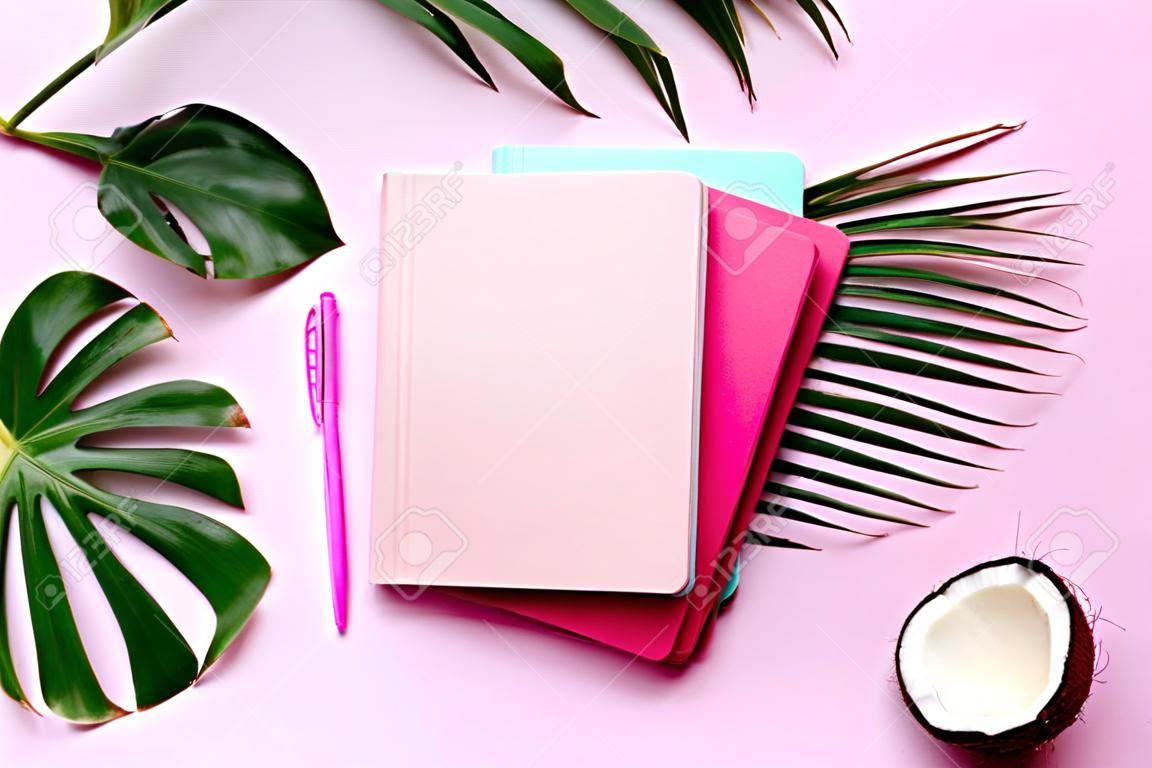 Green monstera, palm leaves, coconut, notebooks over pink background. Female blog writer workspace concept. Flat lay. Top view
