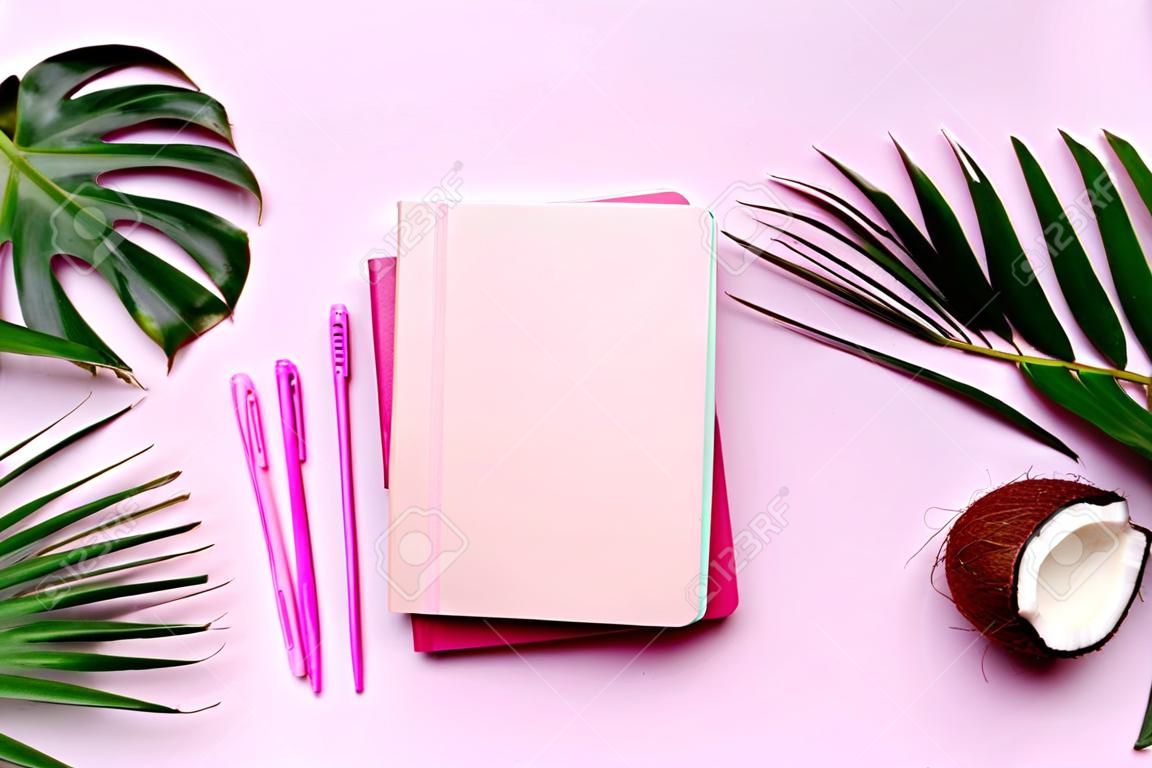 Grüne Monstera, Palmblätter, Kokosnuss, Notizbücher auf rosa Hintergrund. Arbeitsbereichskonzept für weibliche Blogautoren. Flach liegen. Ansicht von oben