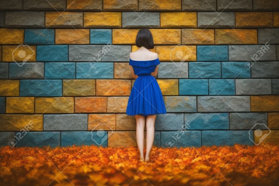 A young beautiful girl in a blue dress standing on a green grass with fallen orange autumn leaves in front of a large stone wall