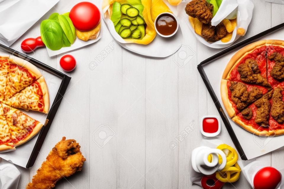 Frame with a variety of take out and fast foods. Pizza, hamburgers, fried chicken and sides. Top view on a white wood background with copy space.